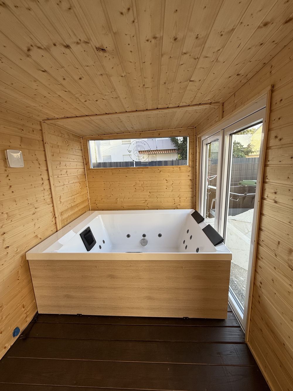 Whirlpool mit Holzverkleidung und zwei Kopfstützen im Wellness Cube, daneben eine Glastür nach draußen.
